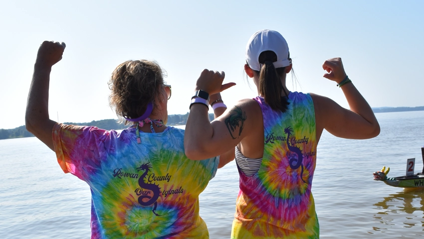 Two women wearing tie dye shirts gesturing with their thumbs to their back while looking away from the camera at High Rock Lake. A dragon boat with the number 2 is just coming into frame on the lower right.